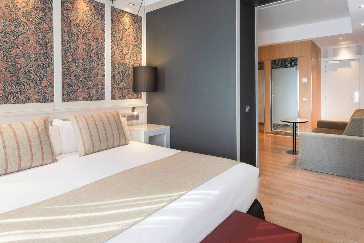 Ein modernes Hotelzimmer mit einem großen Bett, einem Nachttisch, einer Lampe und Blick auf den angrenzenden Wohnbereich.