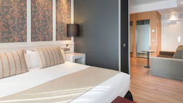 A modern hotel room with a large bed, a bedside table, a lamp, and a view of the adjacent living area.