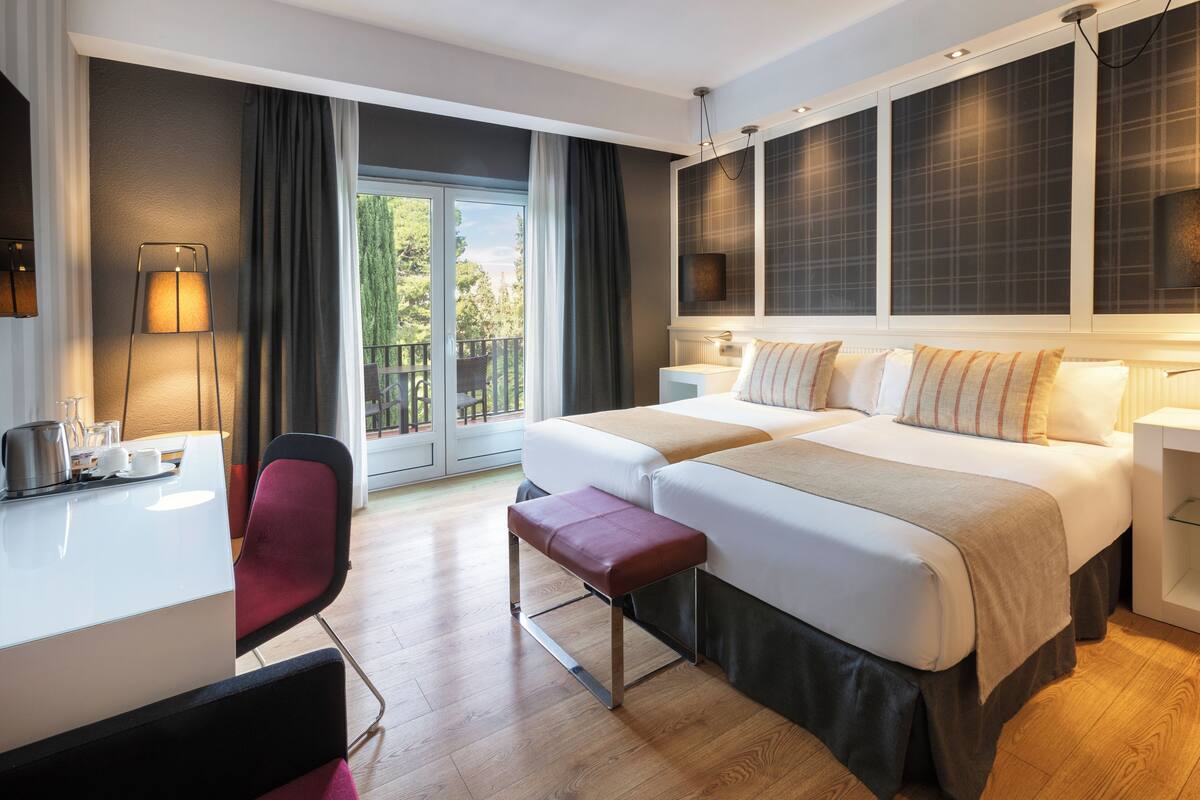 Ein Hotelzimmer mit zwei Betten, einem Schreibtisch, einem Sessel, einer Bank und einem großen Fenster mit Vorhängen.