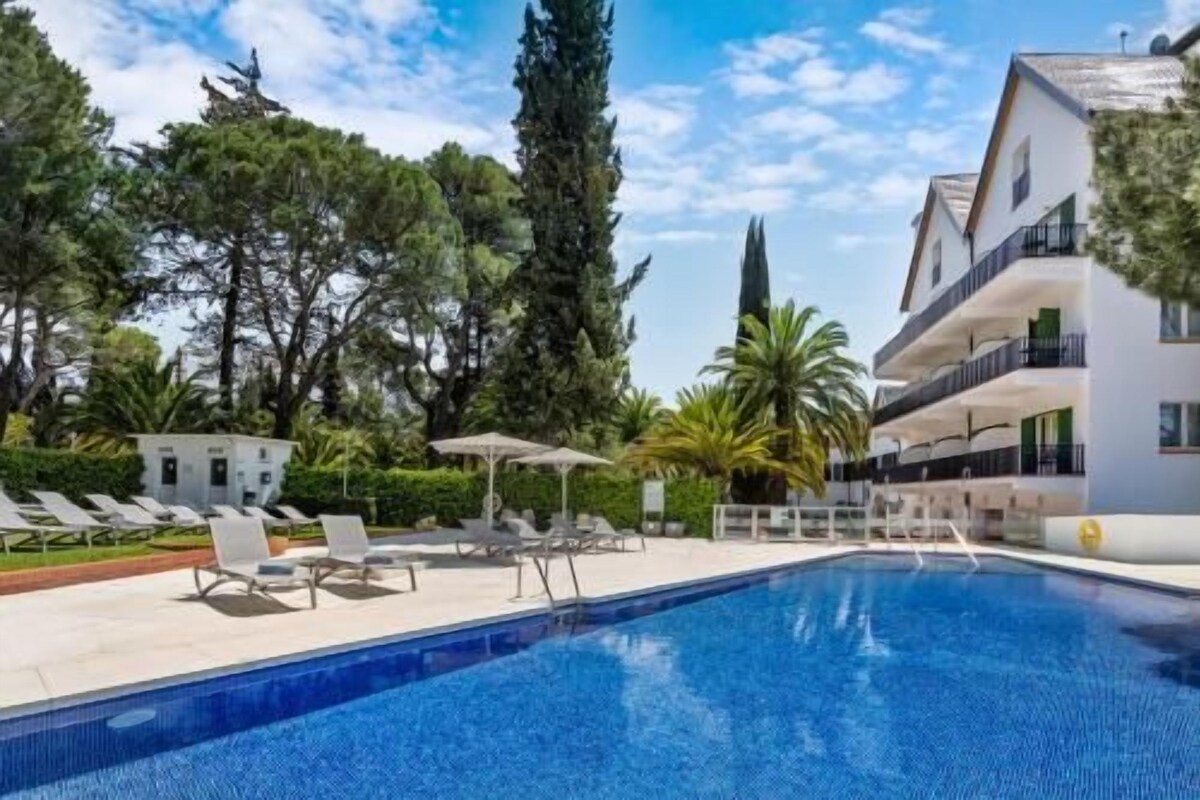 A swimming pool with lounge chairs, a building with balconies, and surrounding trees.