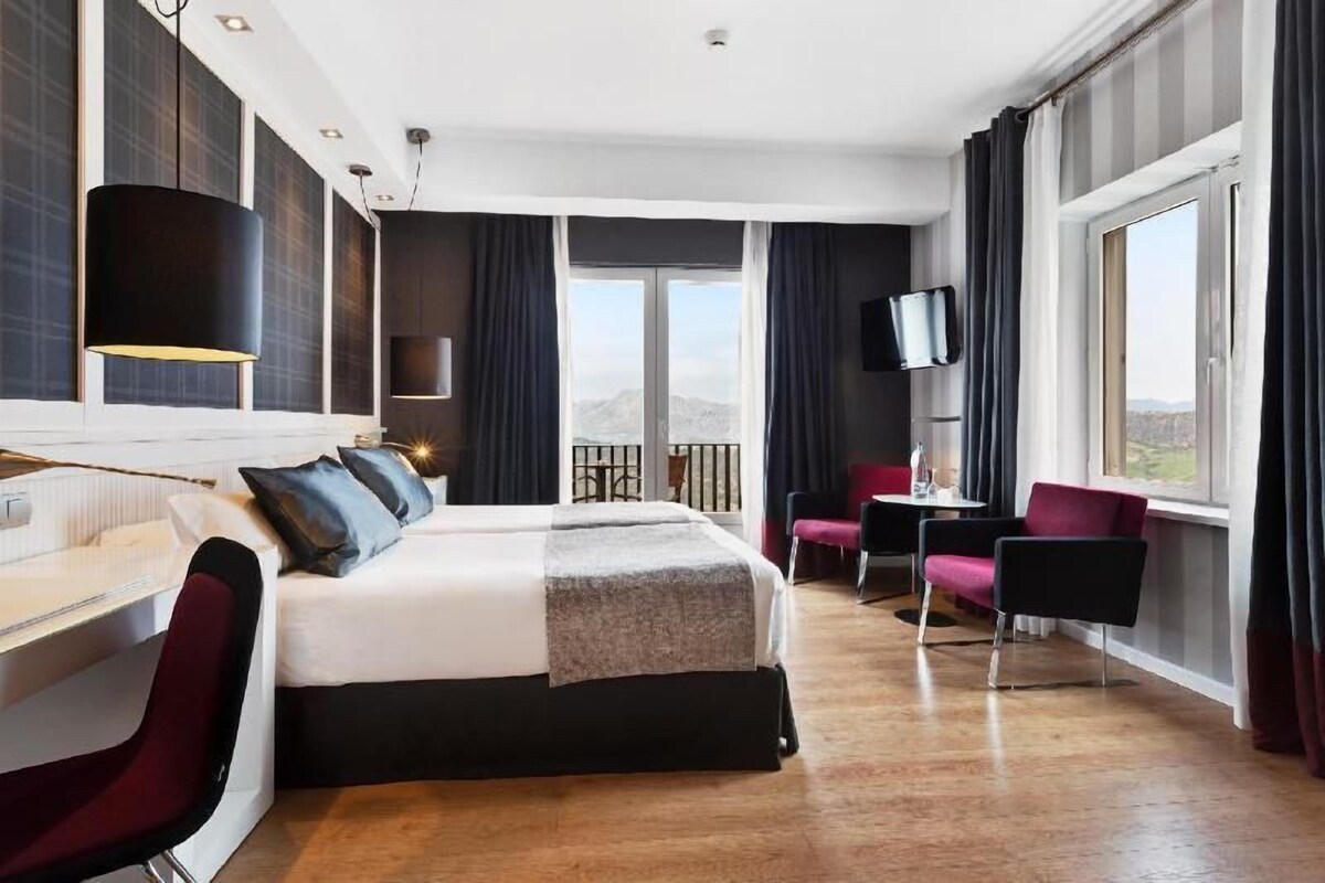 A hotel room with a bed, desk, chairs, a TV, and a balcony with a view.