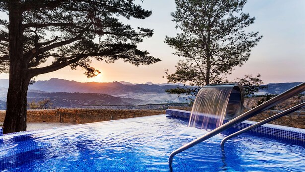 Un jacuzzi con caída de agua, rodeado de árboles y un paisaje montañoso.