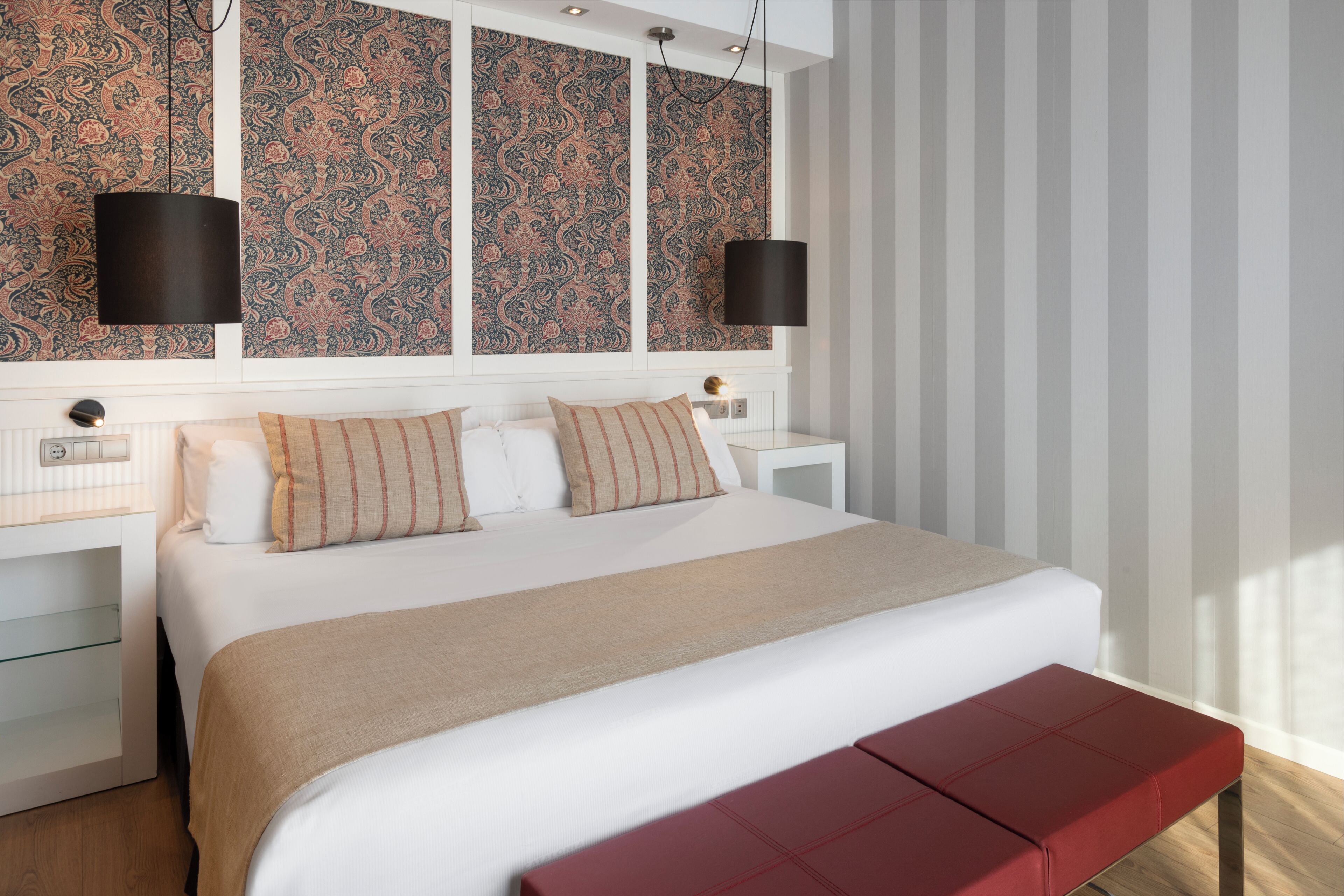 A modern bedroom with a large bed, two bedside tables, a red bench, patterned wallpaper, and two pendant lights.