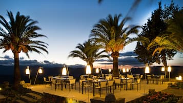 Zona de comedor al aire libre con mesas y sillas, rodeada de palmeras, bajo un cielo vespertino.