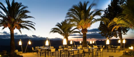An outdoor dining area with tables and chairs, surrounded by palm trees, under evening sky.