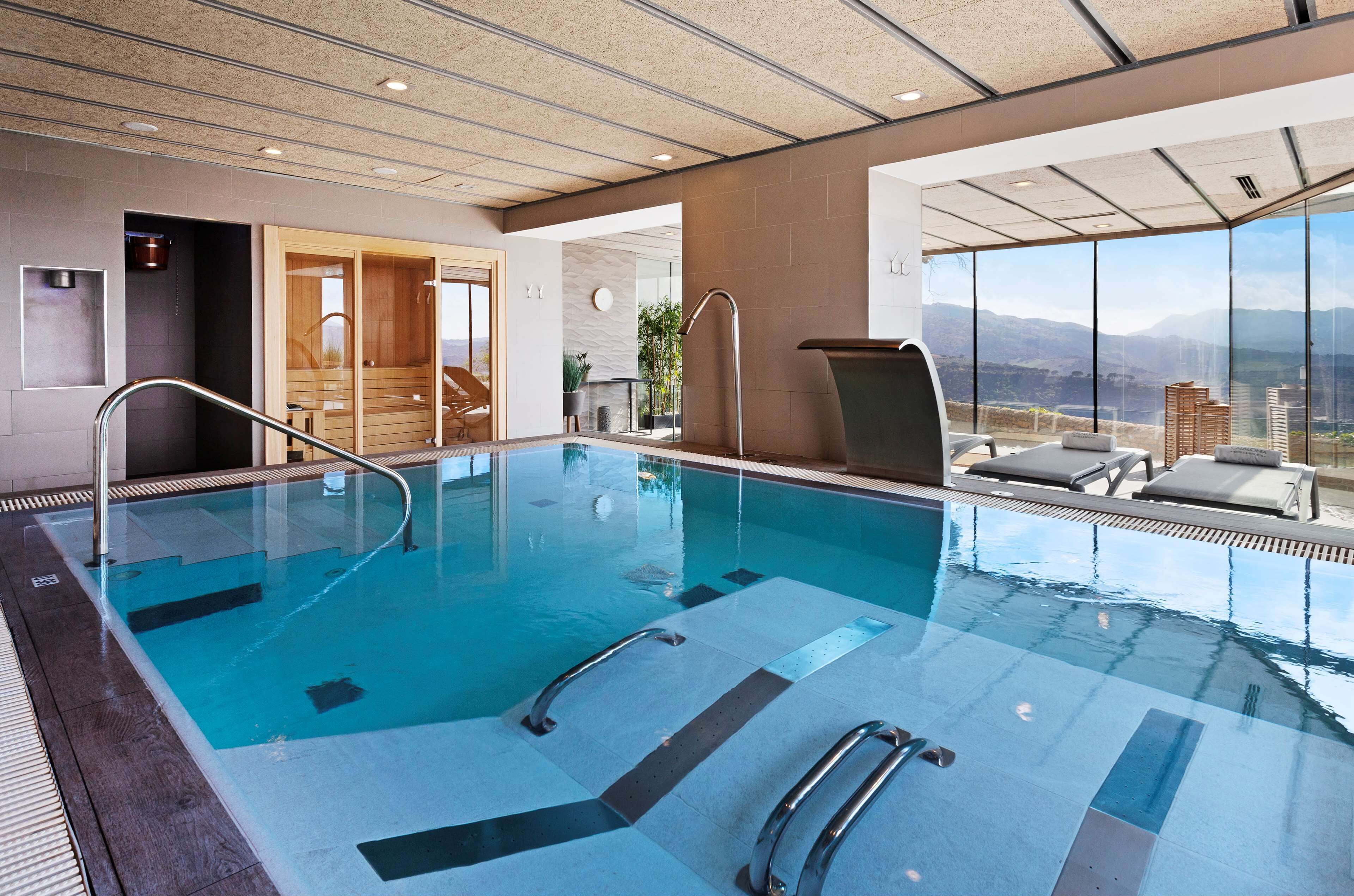 Una piscina interior moderna con paredes de vidrio transparente, vistas a montañas y un área de descanso con sillones reclinables.