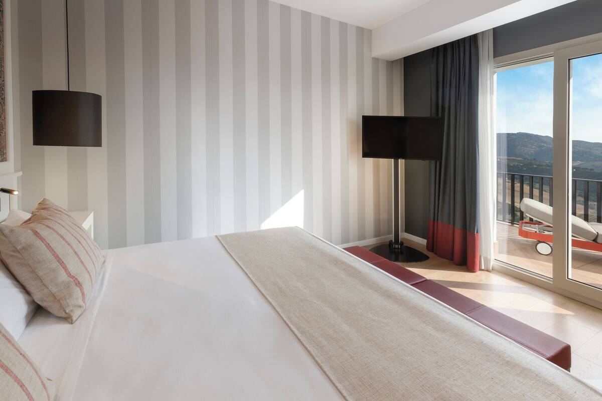 Ein Hotelzimmer mit einem großen Bett, einem Flachbildfernseher, einer Stehlampe und einem Balkon mit Bergblick.