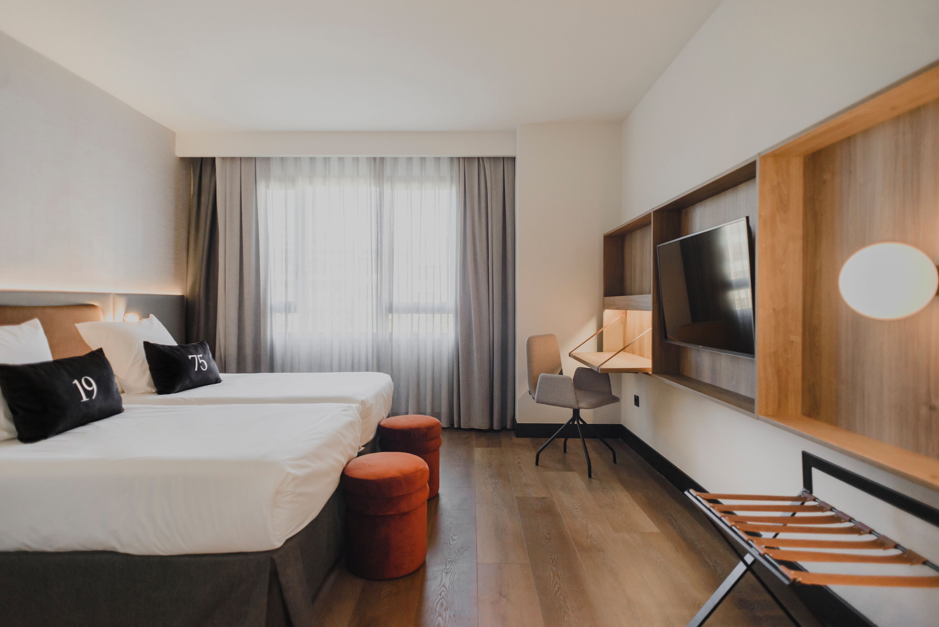 A hotel room with two beds, a desk, a TV, and a large window with curtains.