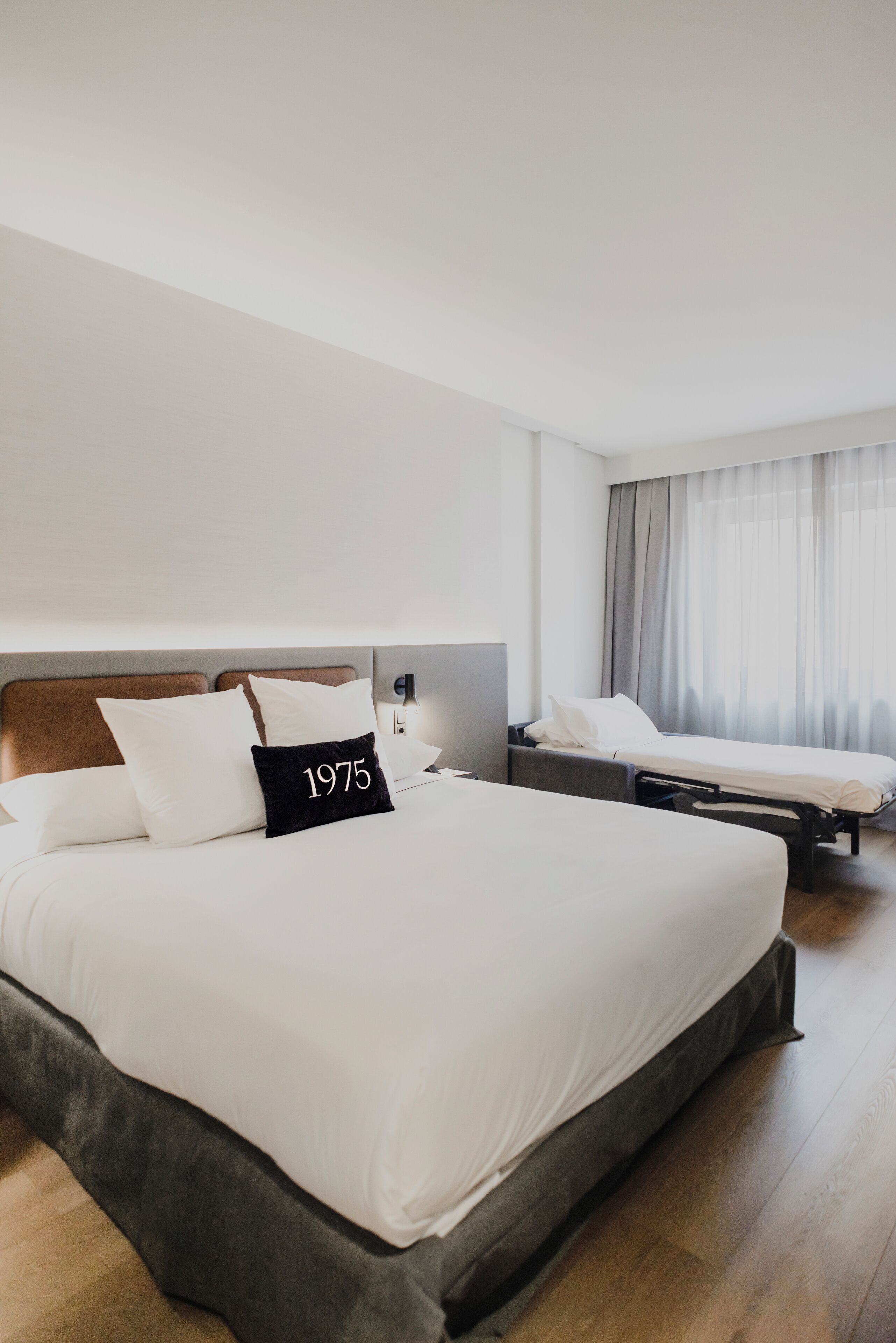 A hotel room with a large bed, a smaller bed, a nightstand, a lamp, and a window with curtains.