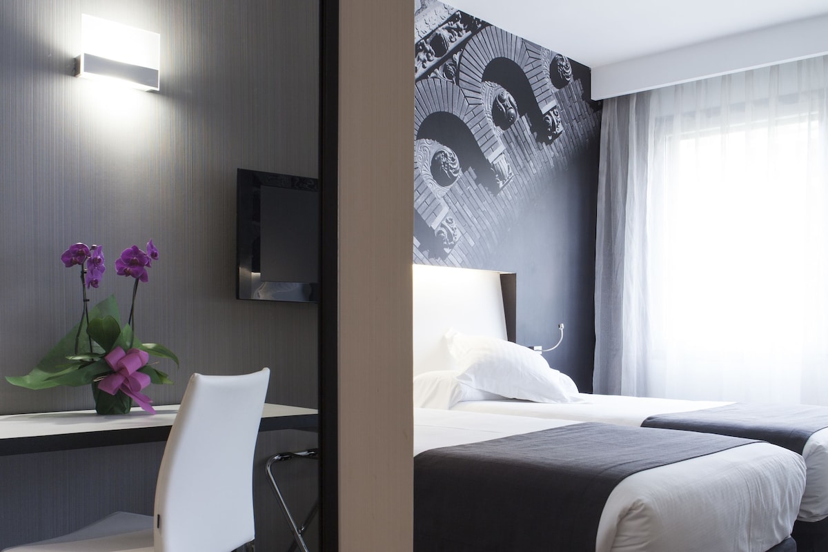 A hotel room with a bed, a desk, a chair, a TV, and a wall with a cityscape mural.