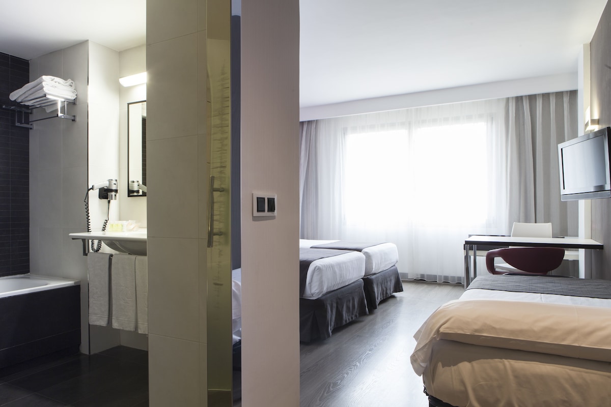 A hotel room with a bathtub, a desk with a chair, two beds, and a TV.