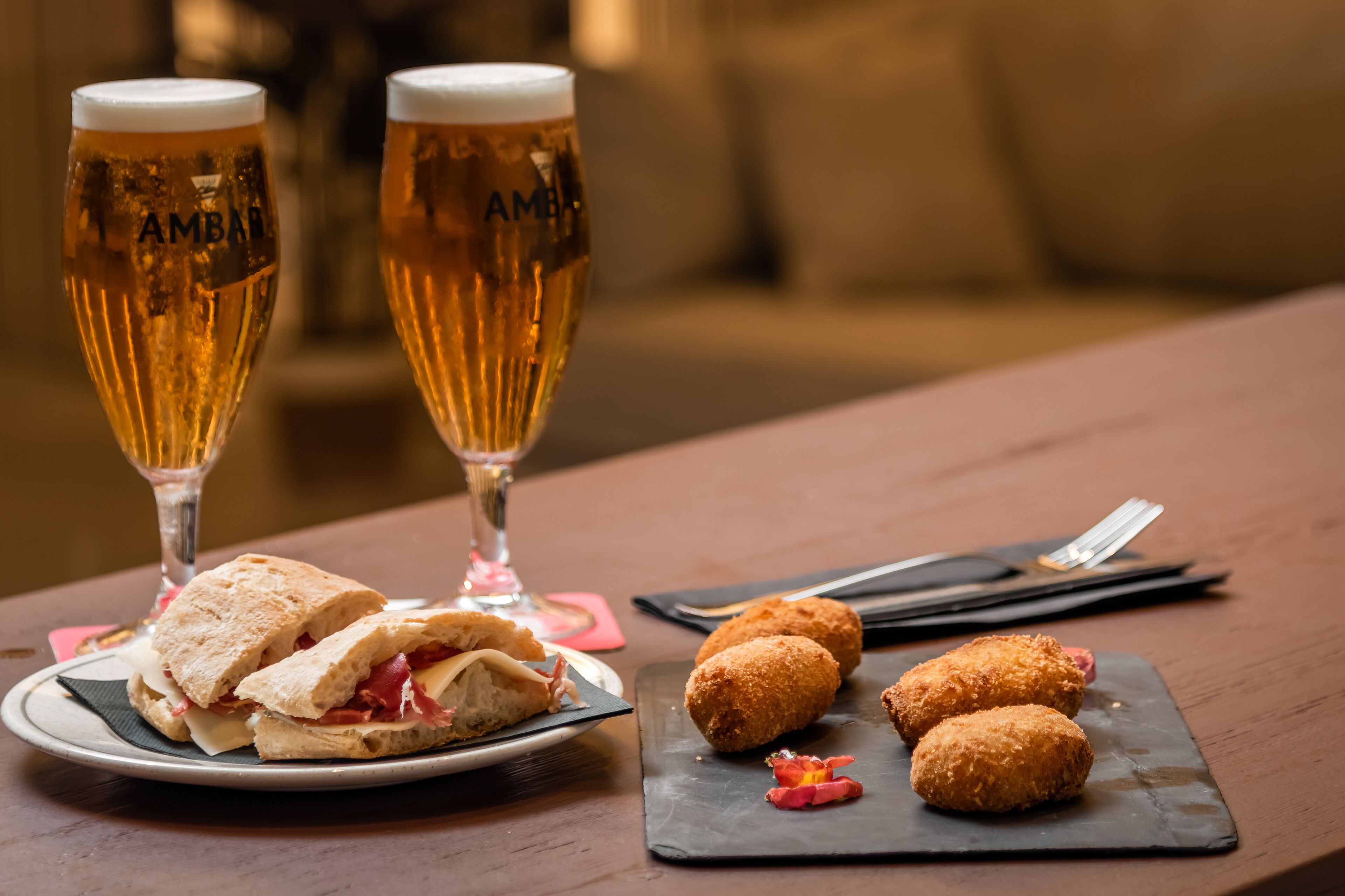 A table with a sandwich, croquettes, and two glasses of beer.