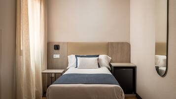 Una habitación de hotel con una cama, mesitas de noche, un armario pequeño, un espejo y una cortina.