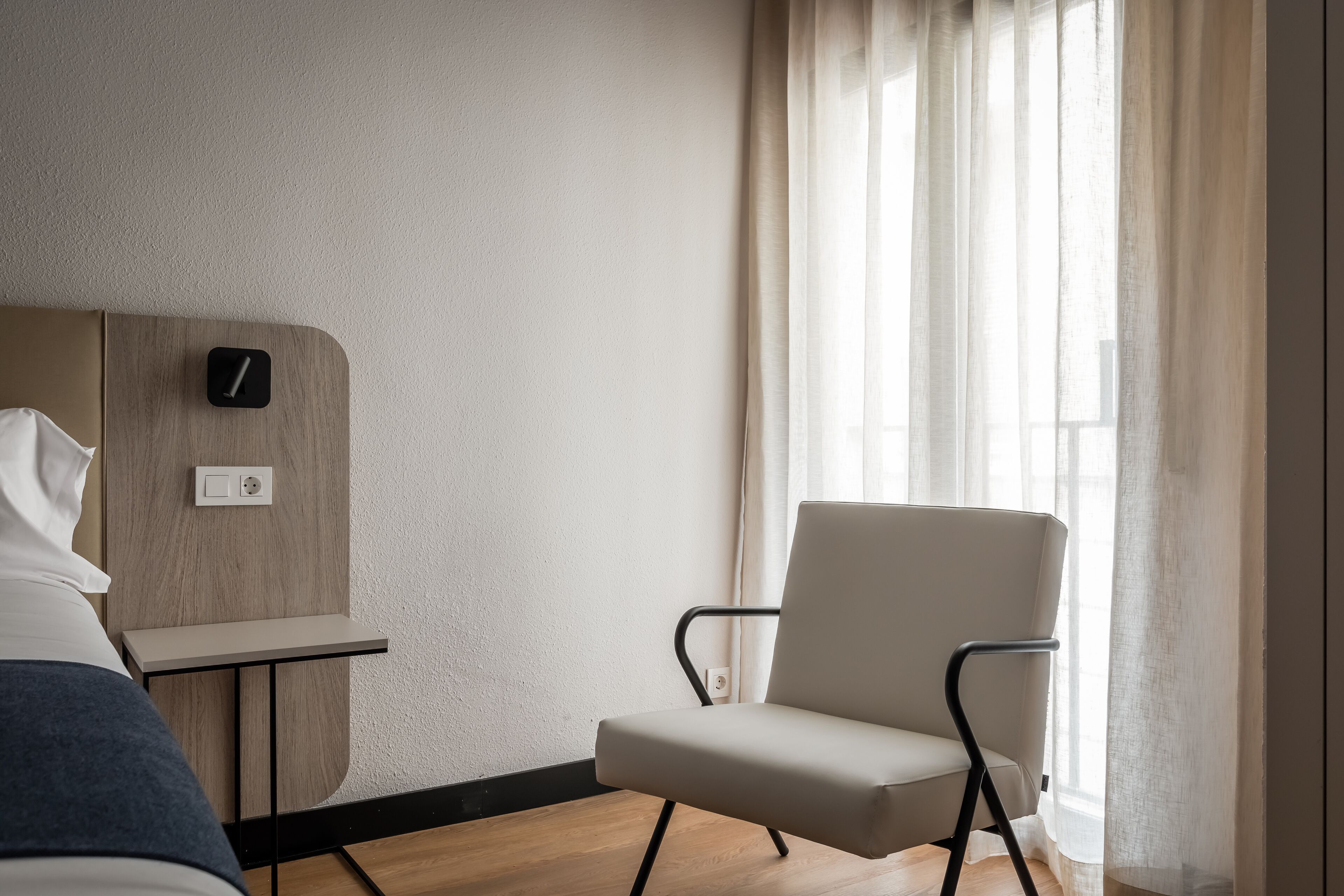 Une chambre d’hôtel moderne, équipée d’un lit, d’une table de chevet, d’une chaise et d’une grande fenêtre avec des rideaux légers.