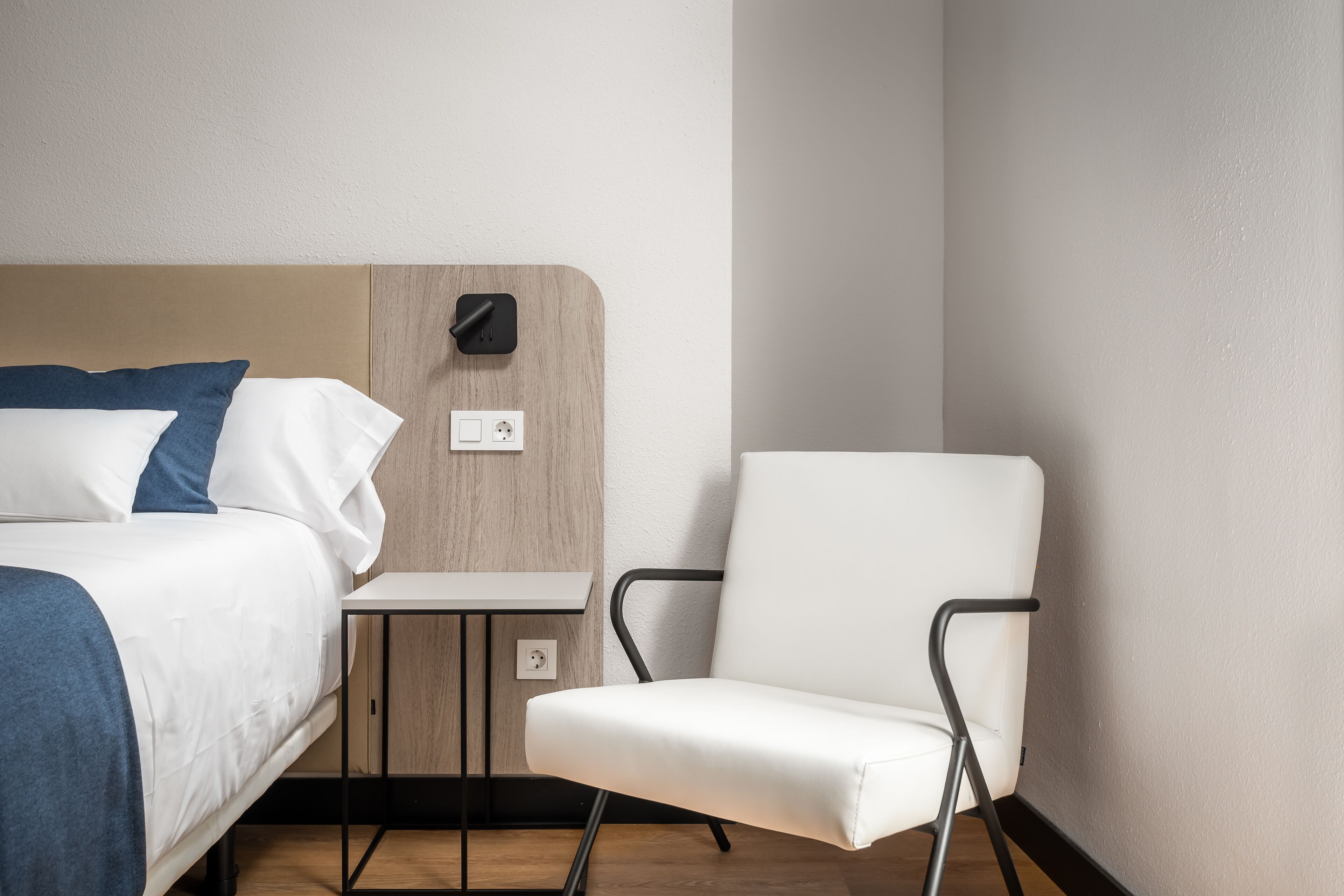 A hotel room with a bed, a chair, a small table, and a wall-mounted light.