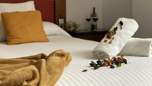 Una cama bien hecha con una manta blanca y marrón, una toalla doblada con flores secas, y una pequeña mesa con una botella de vino al fondo.