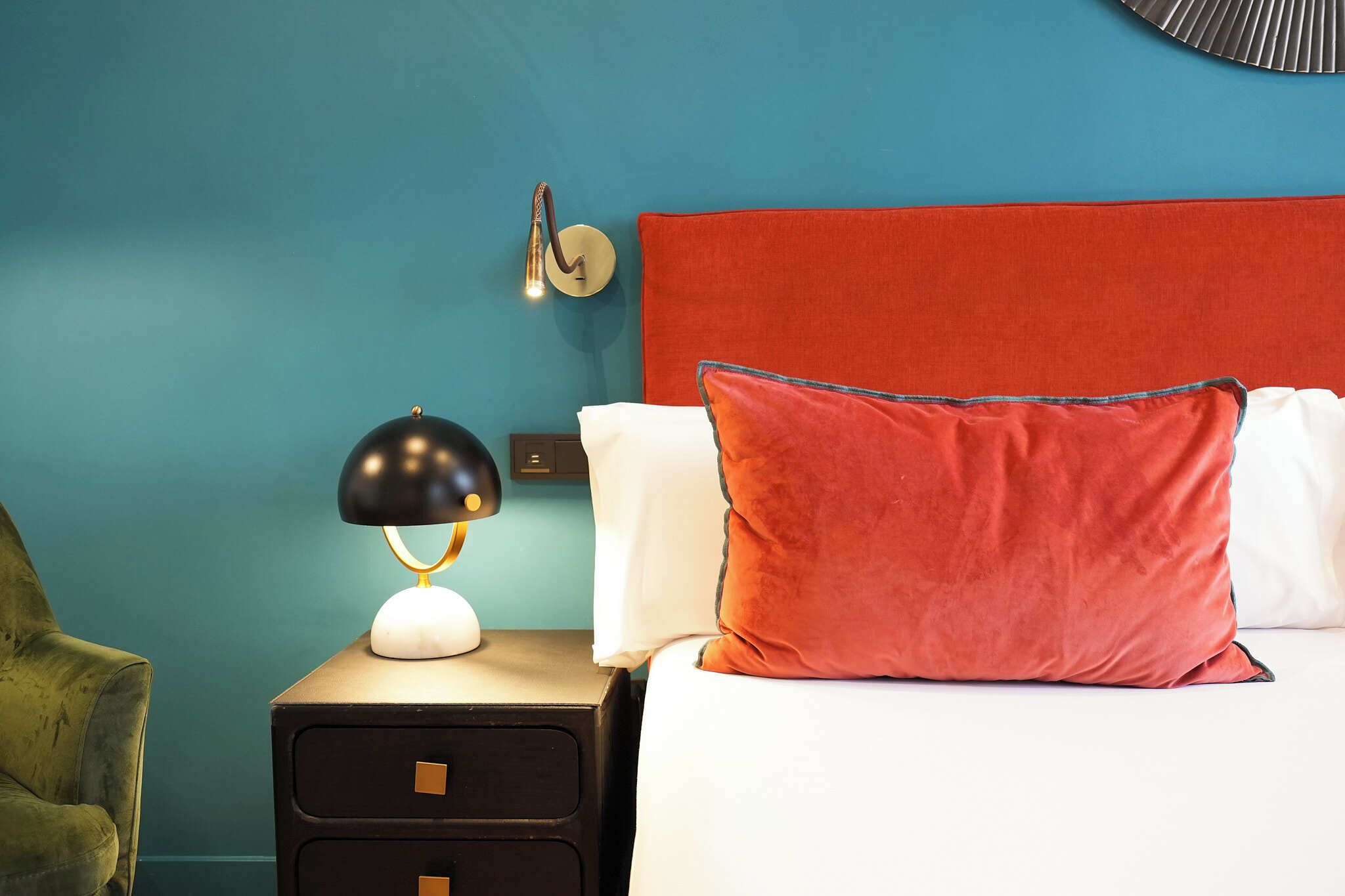 Una cama bien hecha con un cabecero rojo, una almohada blanca y otra roja. Una lámpara negra con base blanca se encuentra sobre una mesita de noche de madera junto a la cama. La pared está pintada de color verde azulado, y hay un reloj de pared redondo encima de la mesita.