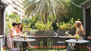 Un patio al aire libre con mesas y sillas redondas, rodeado de follaje y una palmera.