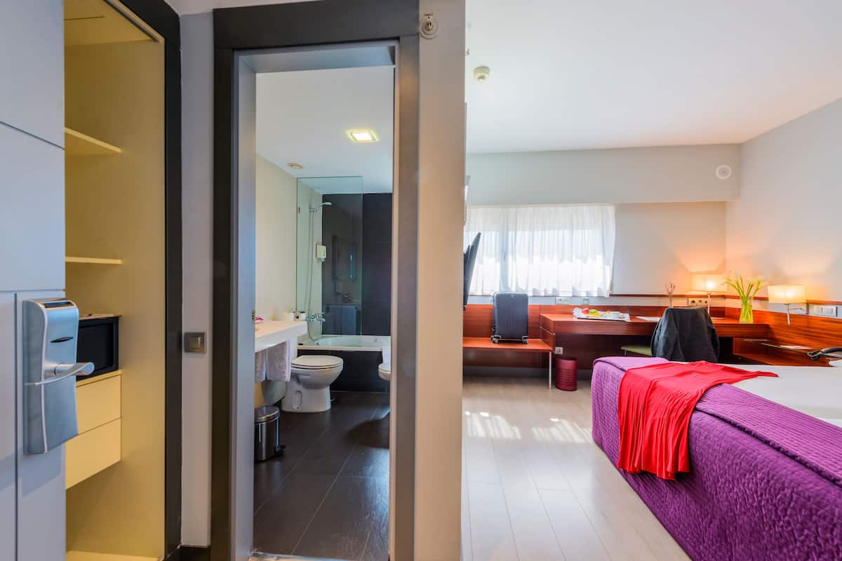 A modern hotel room with a purple bed, a desk, a chair, and a bathroom visible through an open door.