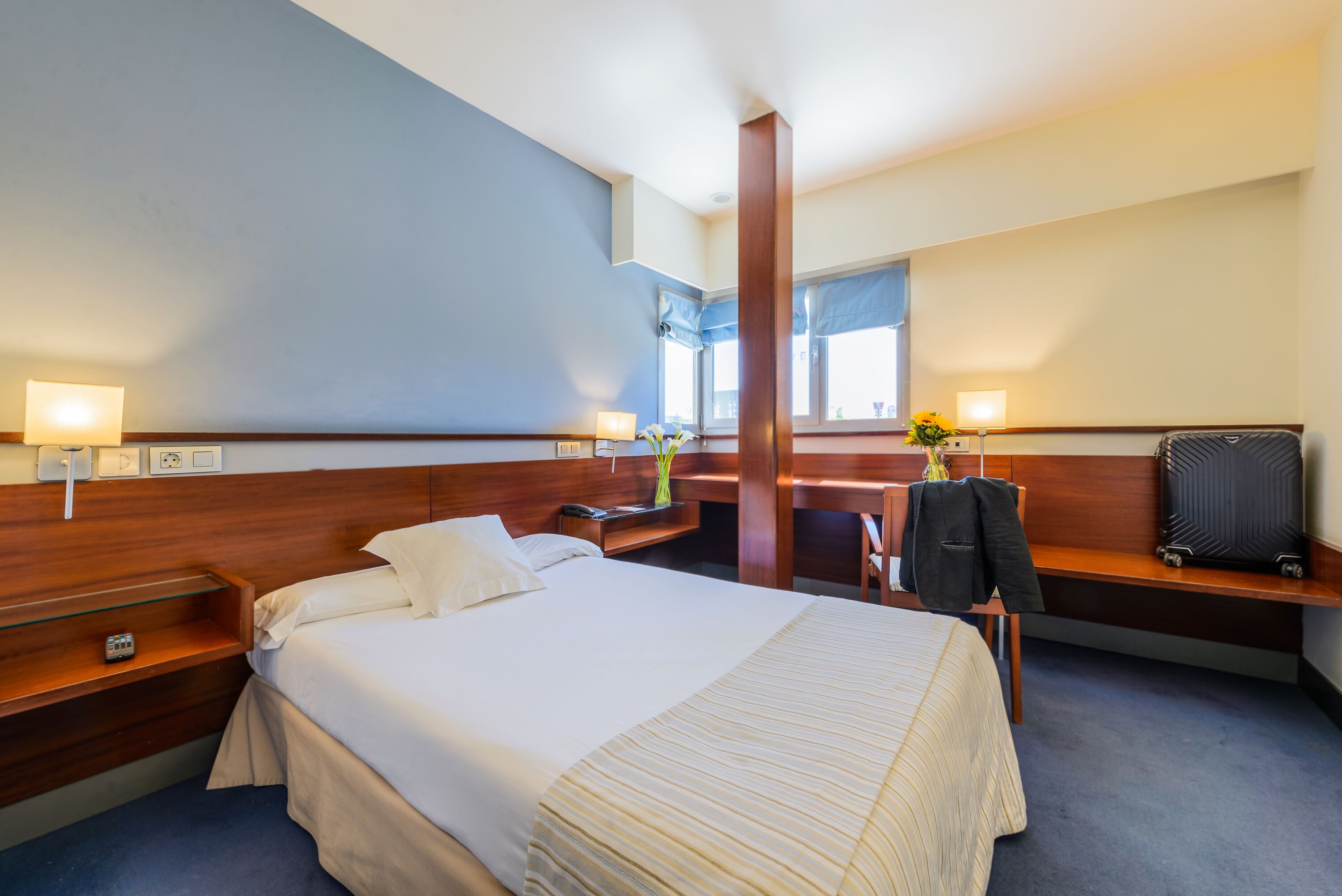 Une chambre d’hôtel avec un grand lit, des tables de chevet, un bureau avec une chaise, un miroir et une fenêtre avec des rideaux.