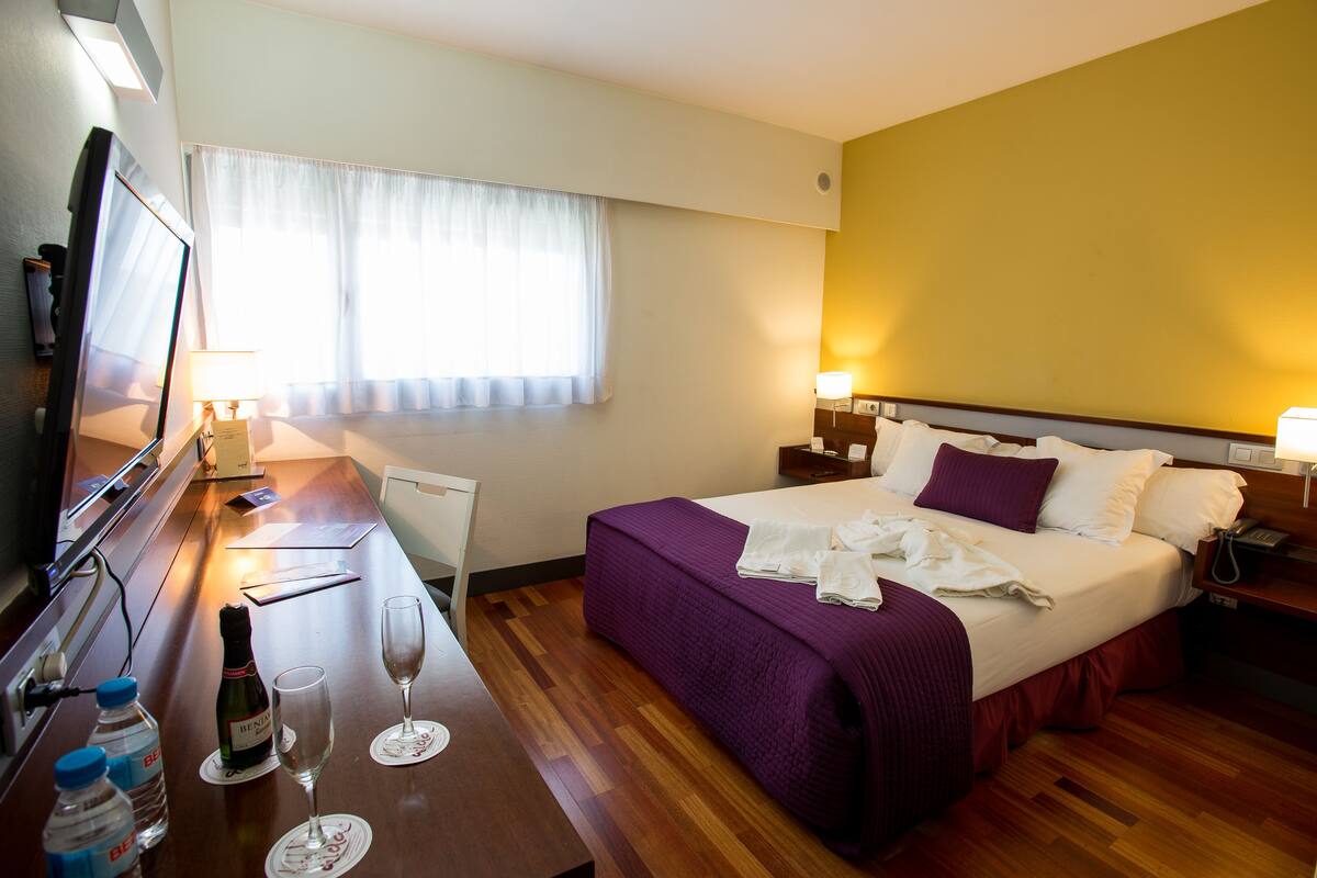 A hotel room with a bed, bedside tables, a desk with a TV, and a window with curtains.