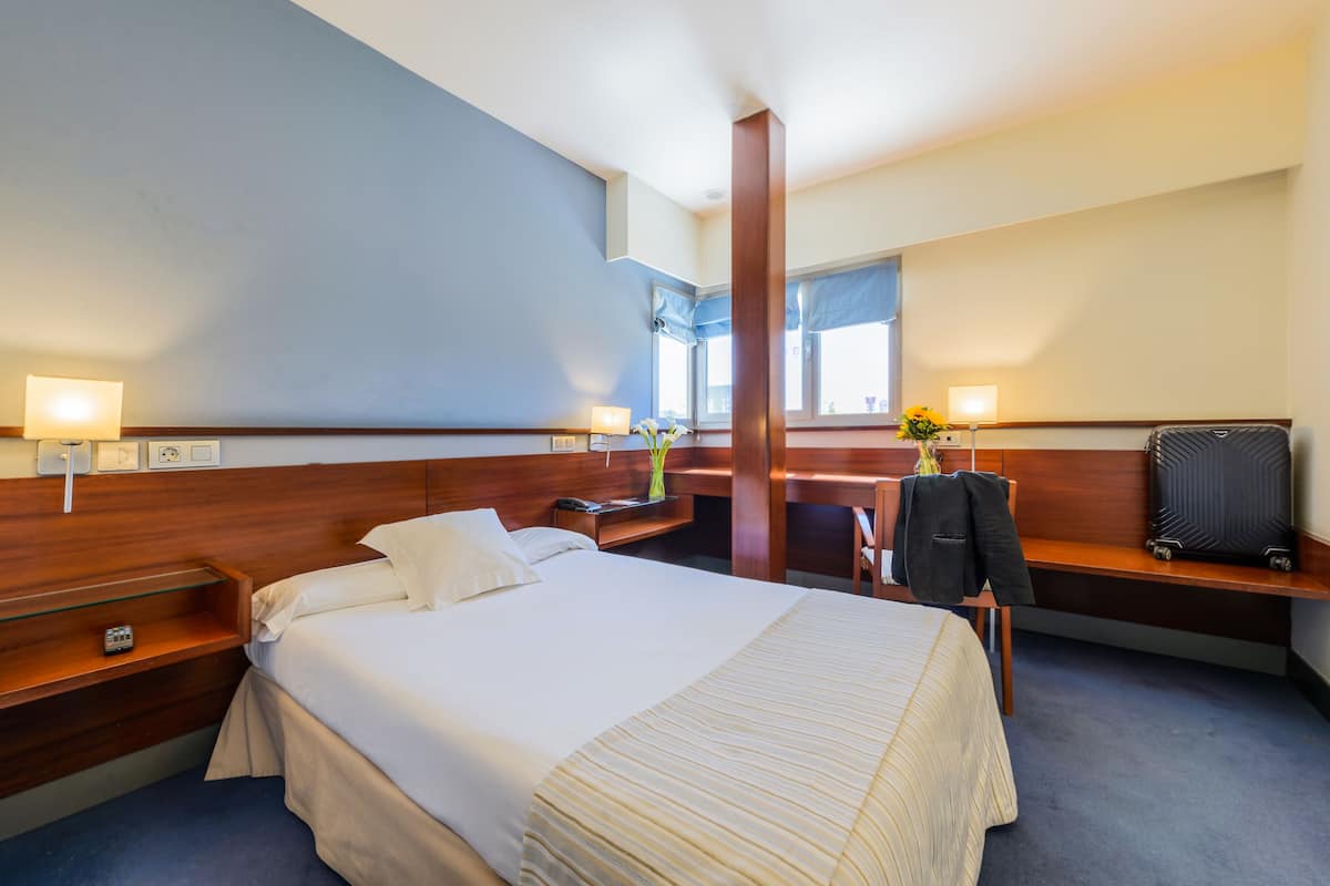 A hotel room with a large bed, bedside tables, a desk with a chair, a mirror, and a window with curtains.
