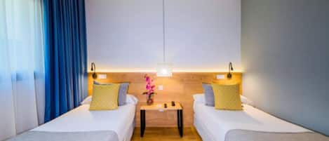 Habitación de hotel con dos camas individuales, una mesita de noche de madera y una pequeña mesa con un florero.