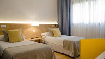Habitación de hotel con dos camas, una mesita de noche, una lámpara y vistas al exterior a través de cortinas.