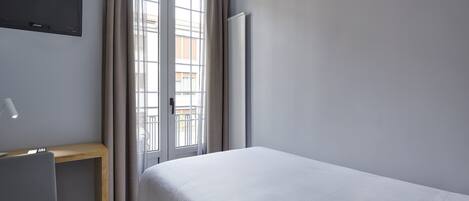 A hotel room with a bed, a desk, a chair, a TV mounted on the wall, and a window with curtains.
