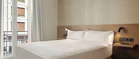 A hotel room with a large bed, a wooden headboard, a nightstand, and a wall-mounted lamp.