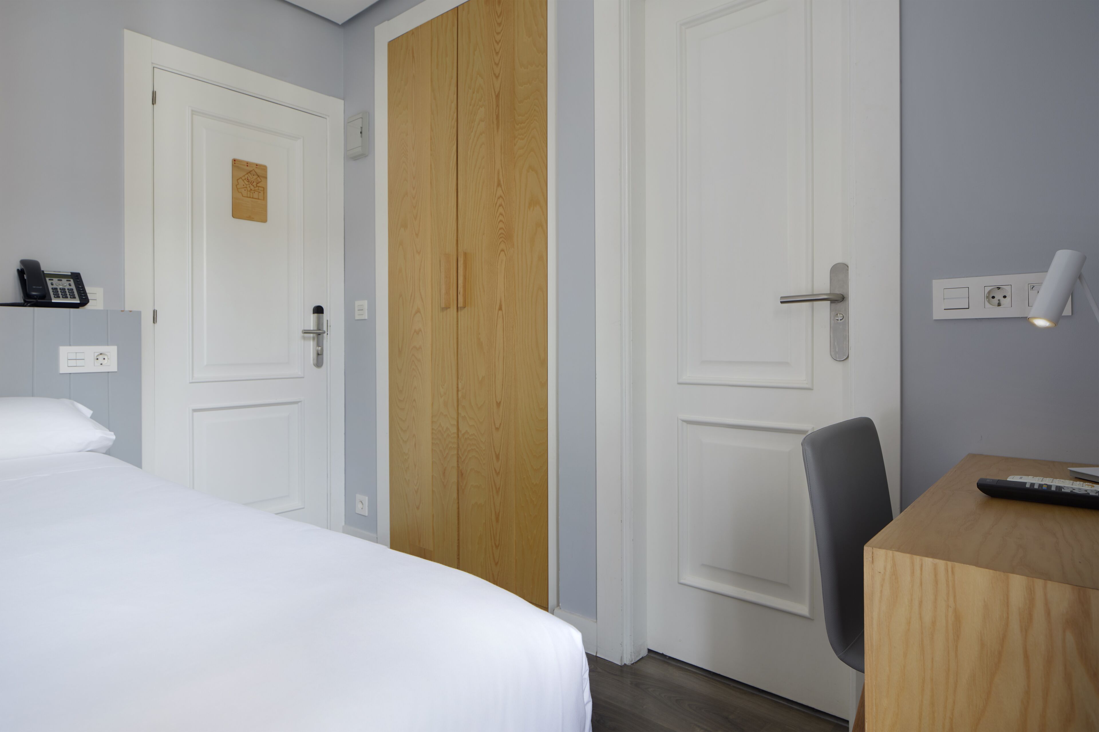 Habitación de hotel con cama, armario de madera, escritorio con silla y una lámpara en la pared.