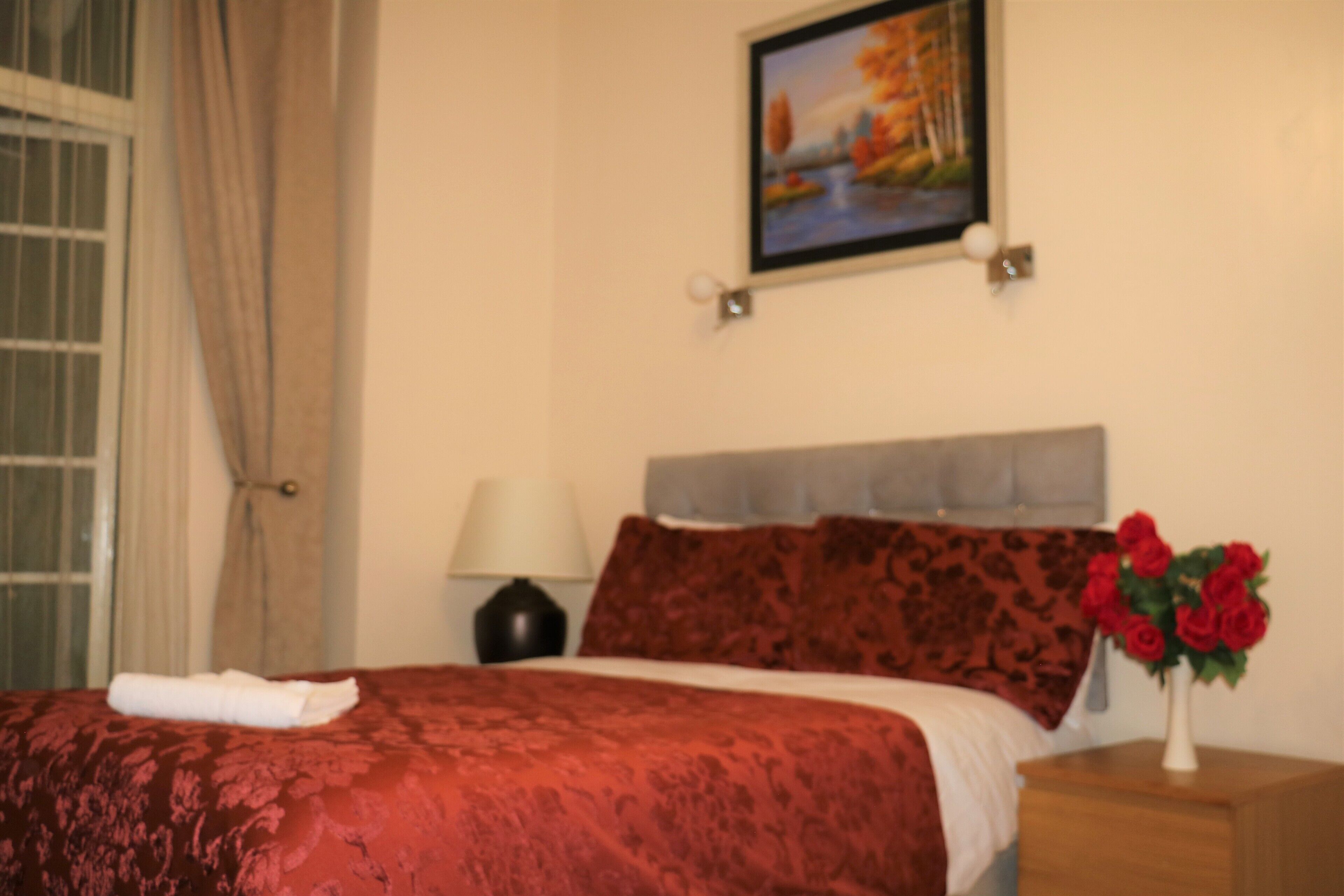 A hotel room with a bed, a nightstand, a lamp, a painting on the wall, and a vase with flowers.