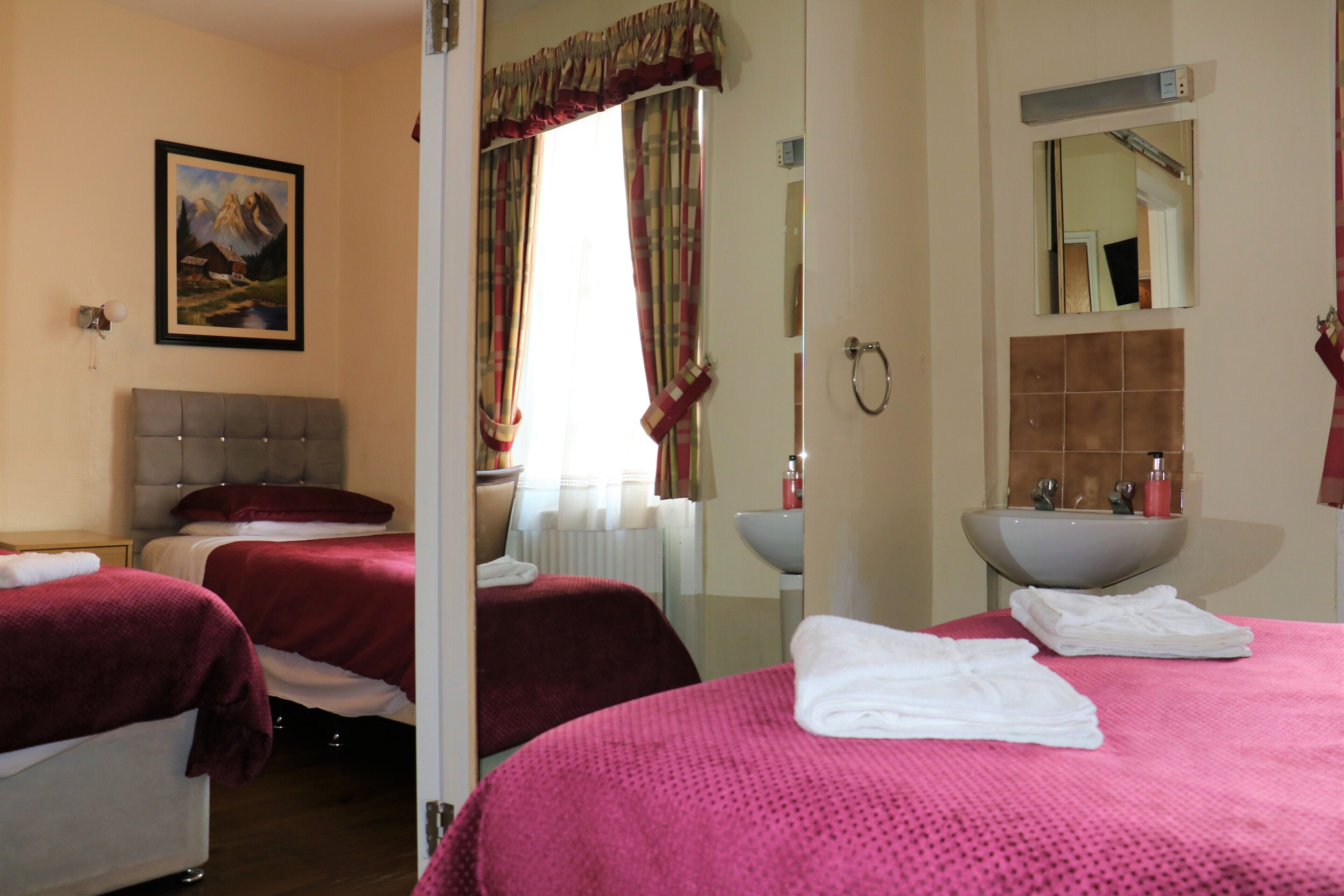 A hotel room with two beds, a large window with curtains, a bathroom with a sink and mirror, and a painting on the wall.