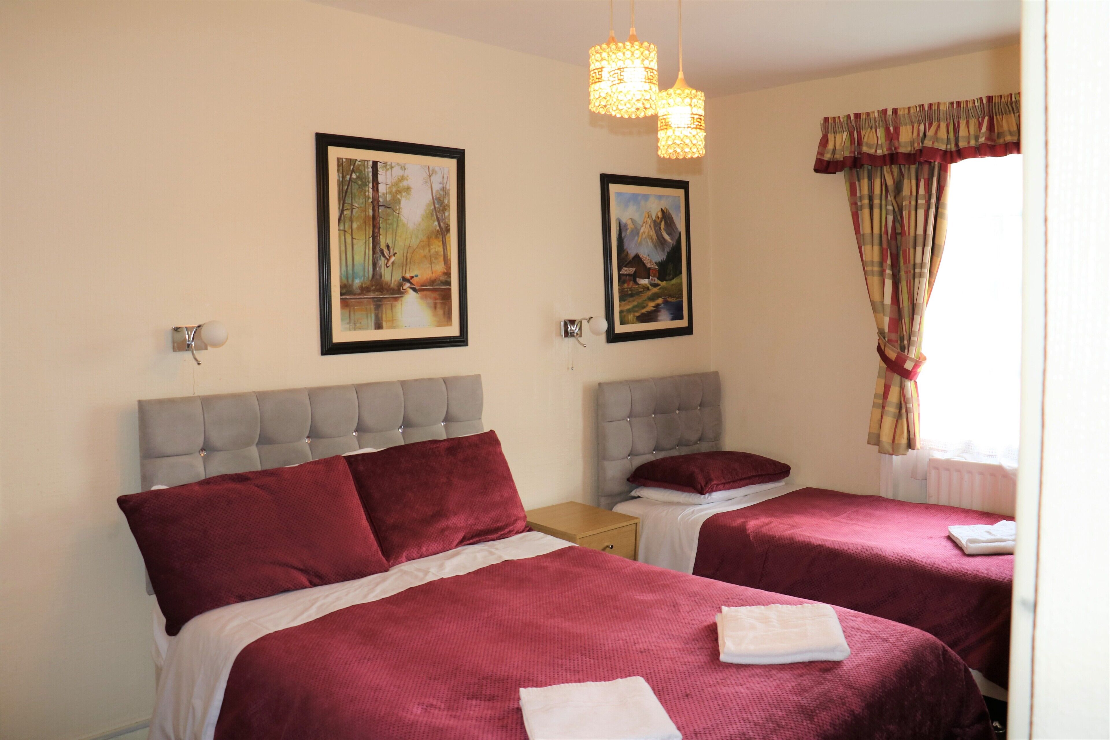 Habitación de hotel con dos camas, cada una con cabecero gris y ropa de cama roja. Hay dos cuadros enmarcados en la pared, una ventana con cortinas y una lámpara colgante.