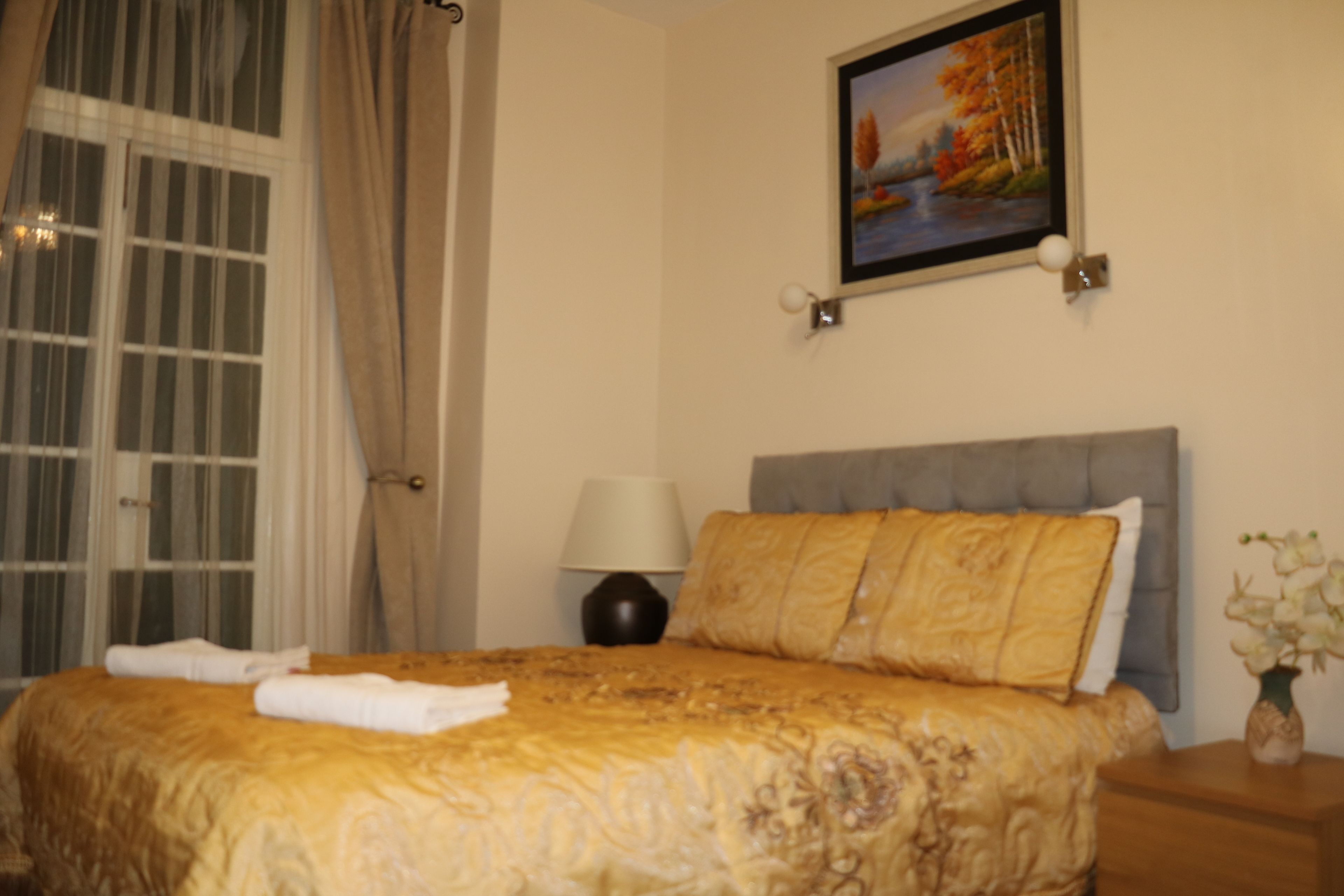 Een hotelkamer met een bed, nachtkastje, lamp, schilderij en een raam met gordijnen.