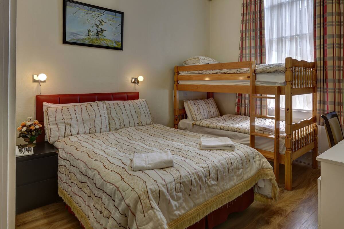 A room with a bunk bed, a bedside table with a lamp, a small desk with a kettle, and a window with curtains.