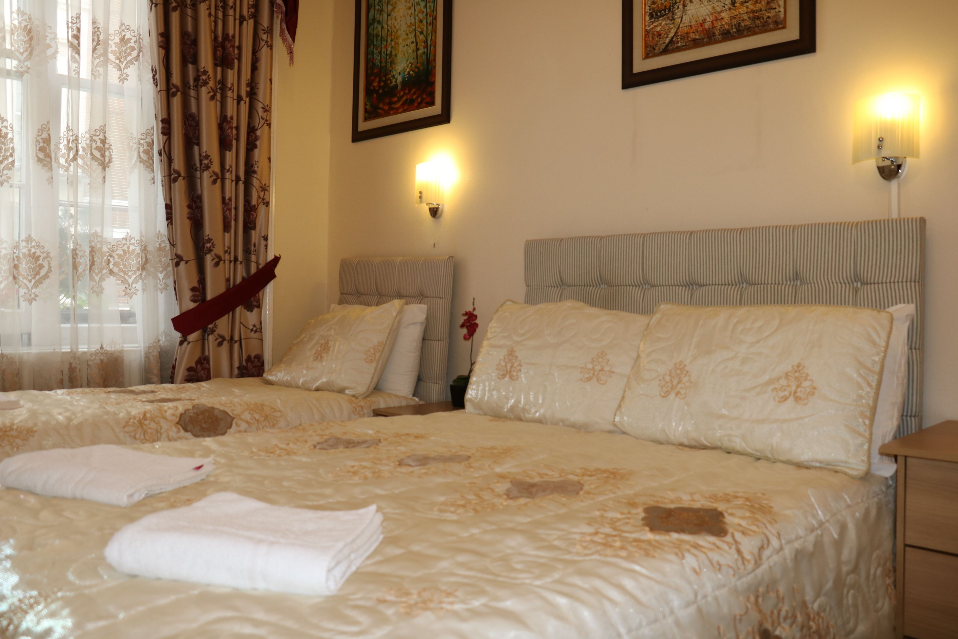 A hotel room with two beds, each with a headboard and multiple pillows. There are wall-mounted lamps and a painting on the wall.