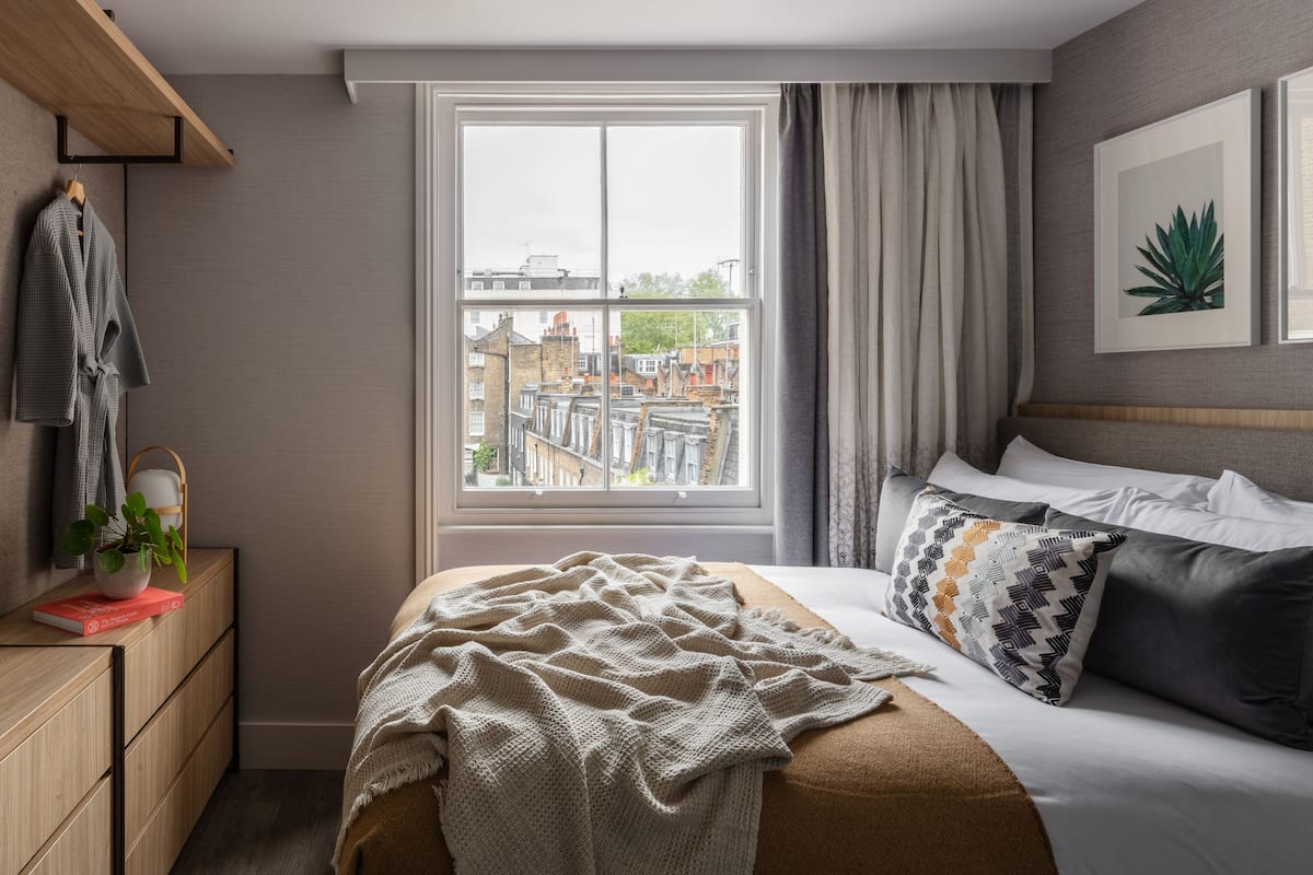 A bedroom with a large window, a bed with patterned pillows, a bedside table with a plant, and a robe hanging on the wall.