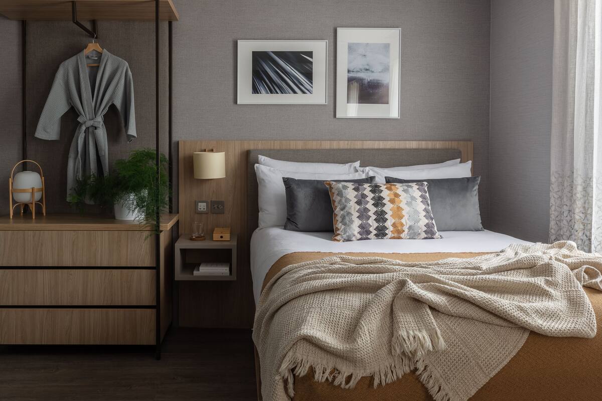 A modern bedroom with a wooden headboard, a bedside table with a lamp, a robe hanging on a rack, and a bed with multiple pillows.