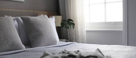 A neatly made bed with a grey blanket and pillows, a bedside lamp, and a potted plant by a window with curtains.