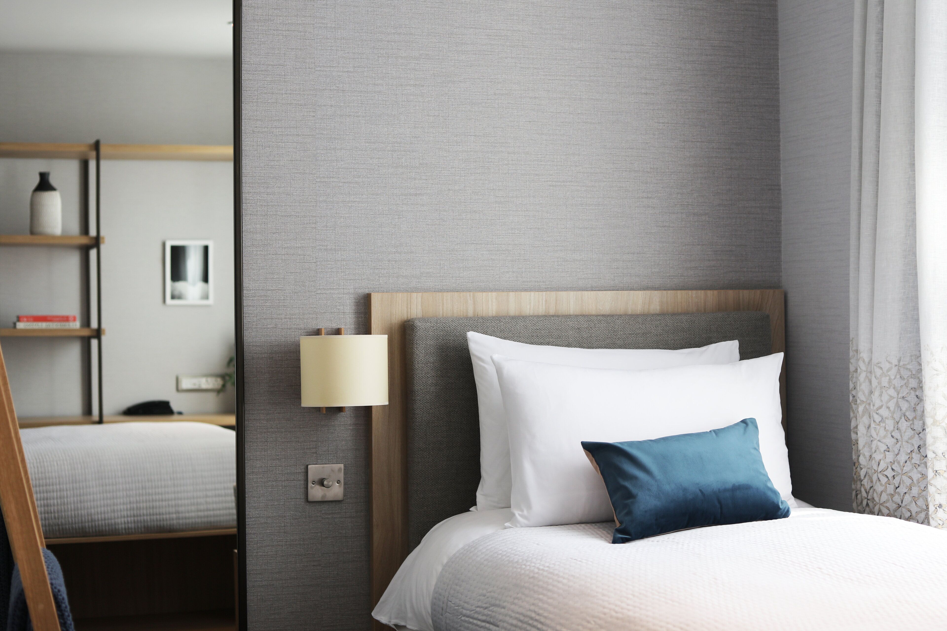 A hotel room with a bed, a nightstand, a lamp, and a shelf with books and a vase.