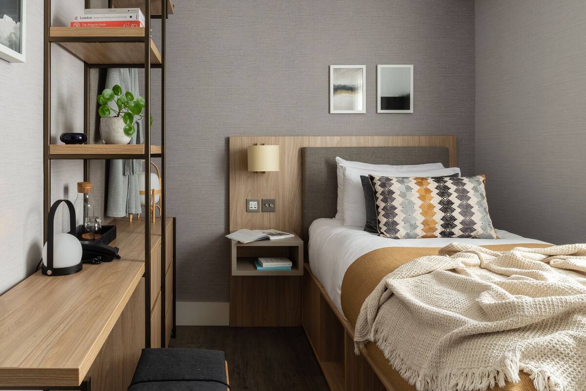 A modern hotel room with a wooden bed, a bedside table, a desk, and a bookshelf.