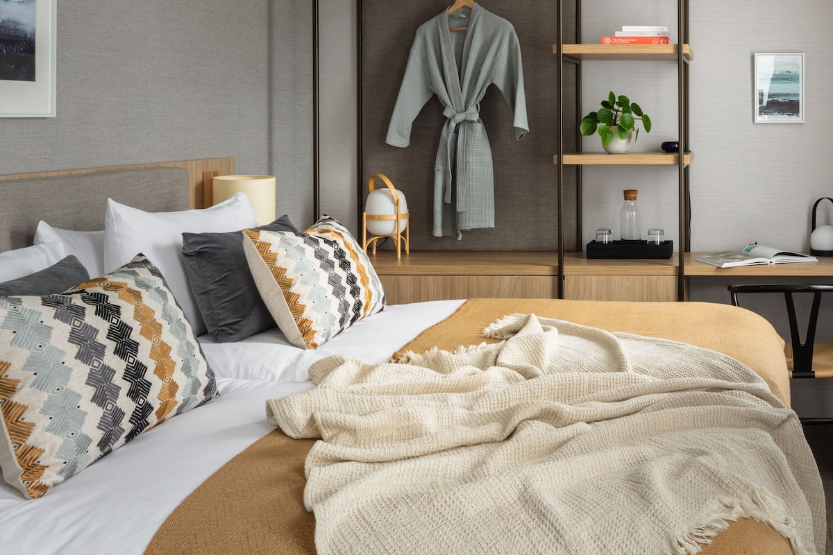 A modern bedroom with a bed, pillows, a bedside table, a nightstand, a robe, a shelf with books, a plant, and a framed picture on the wall.
