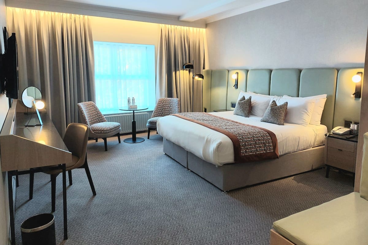 A hotel room with a large bed, a desk, a chair, a small table, and a TV.