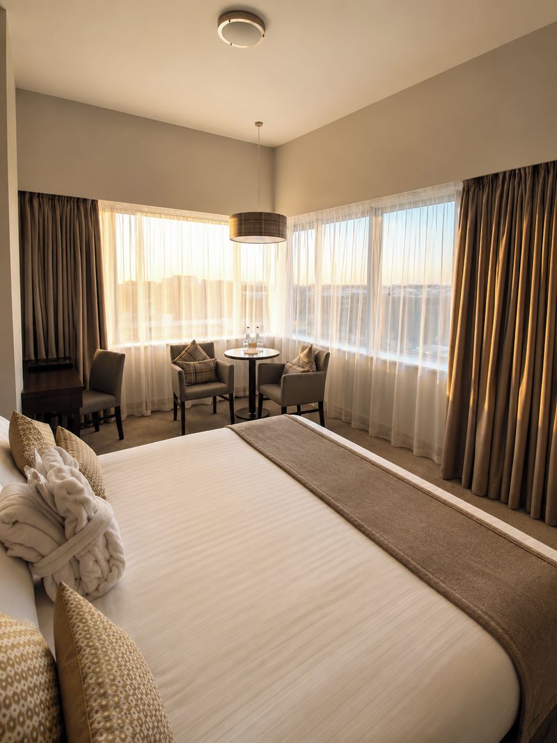 Quarto de hotel com uma cama grande, área de estar com cadeiras, uma mesa pequena e vista pela janela.