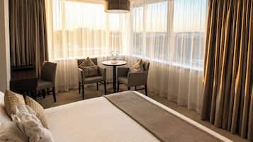 Quarto de hotel com uma cama grande, área de estar com cadeiras, uma mesa pequena e vista pela janela.