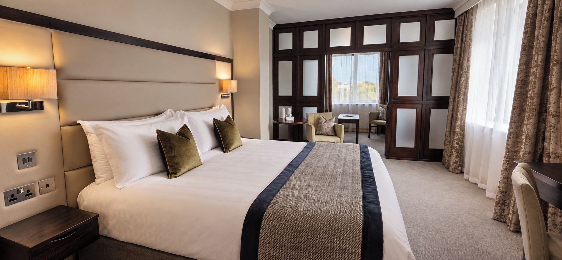 A hotel room with a large bed, bedside lamps, a small table, and a view through a window with curtains.