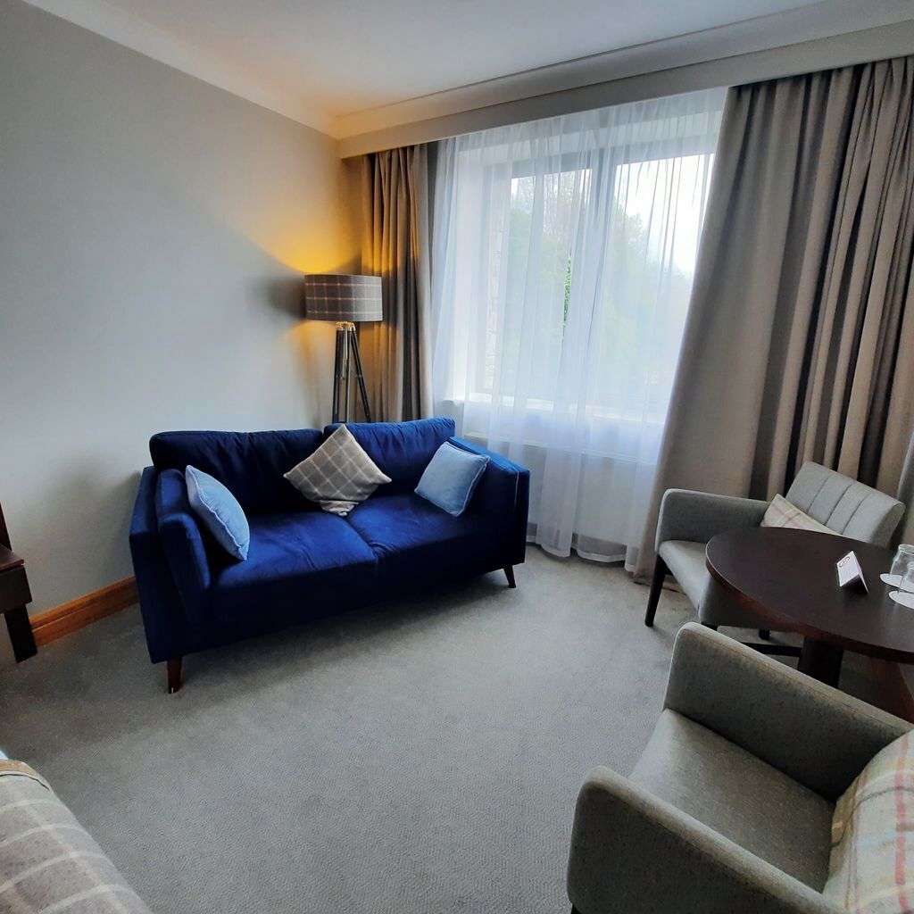 A hotel room with a blue sofa, a grey armchair, a small table with a name card, and a floor lamp.