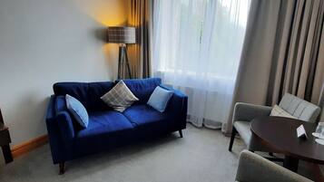 A hotel room with a blue sofa, a grey armchair, a small table with a name card, and a floor lamp.