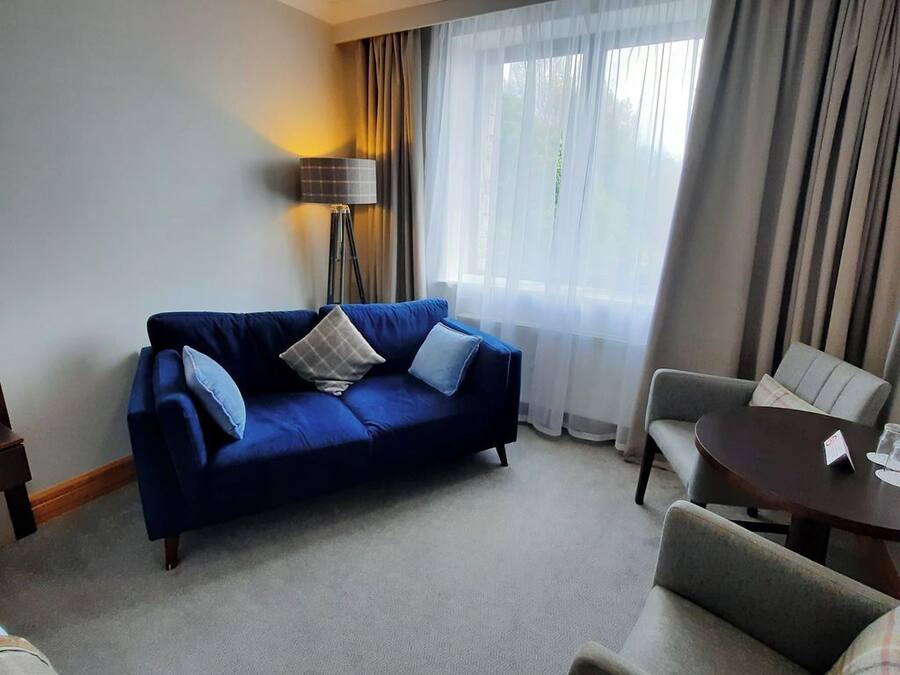 A hotel room with a blue sofa, a grey armchair, a small table with a name card, and a floor lamp.